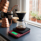 Coffee scale with timer and backlit display under a pour-over brew on a sunlit kitchen counter