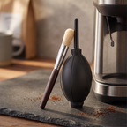 Grinder cleaning brush and air blower bulb on a slate counter beside a coffee grinder