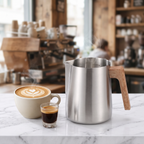 Stainless latte art pitcher with wood-grain handle on marble cafe bar beside heart latte