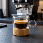 Double wall glass espresso cup on a bamboo base holding fresh espresso on a slate counter