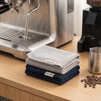 Barista microfiber cleaning towels stacked on a light oak coffee bar beside an espresso machine steam wand